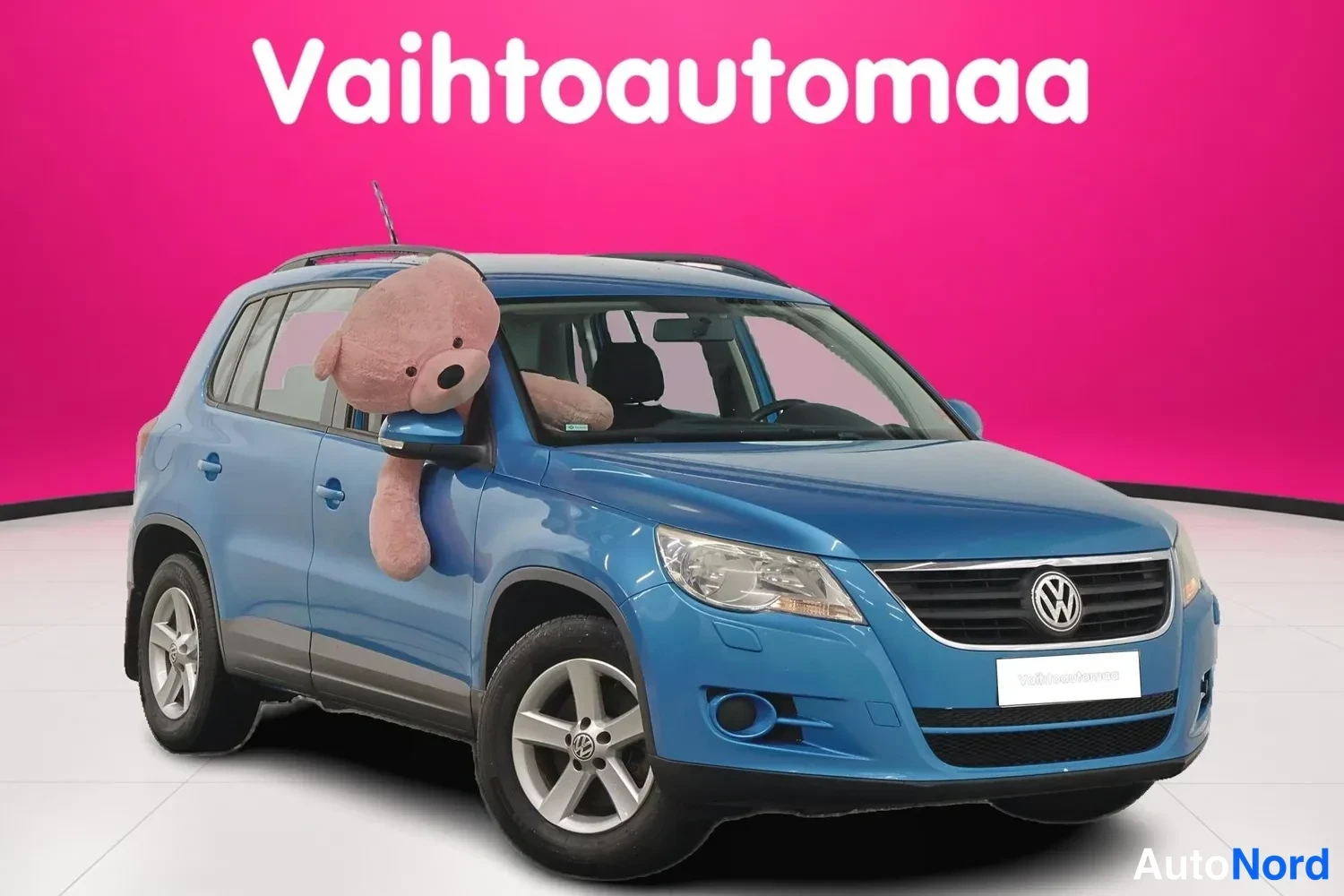 Etuviisto, nokka oikealle: Sininen tila-auto kolmivartalokuvakulmassa etuosa oikealle, pehmoeläin tuulilasin kohdalla, vaaleanpunainen tausta.