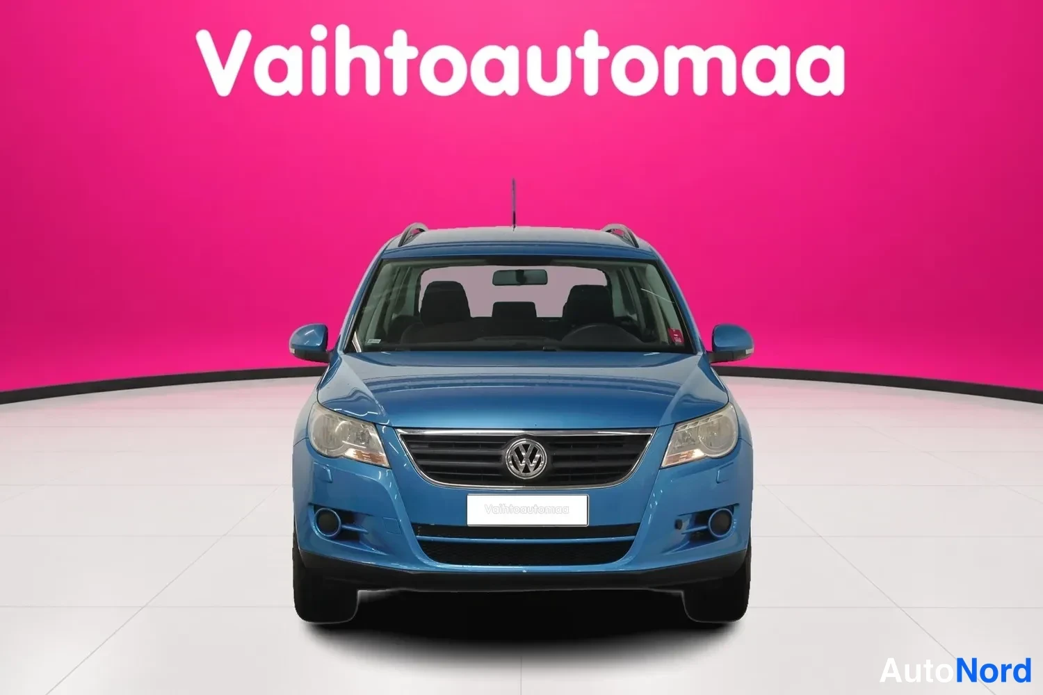 Edestä: Sininen tila-auto suoraan edestä kuvattuna, vaaleanpunainen tausta ja vaalea lattia.