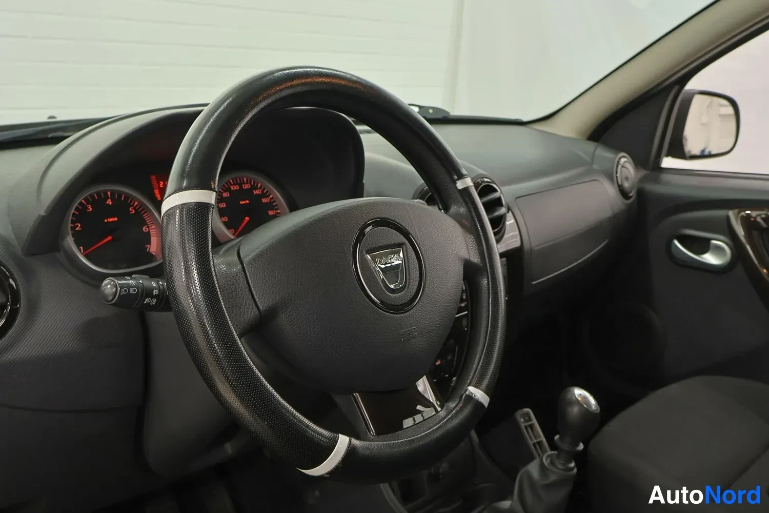 Muu: Ohjauspyörä Dacia-merkinnällä ja mittaristo punaisin neuloin kuvattuna, käsivaihteen vipu näkyvissä keskikonsolissa.