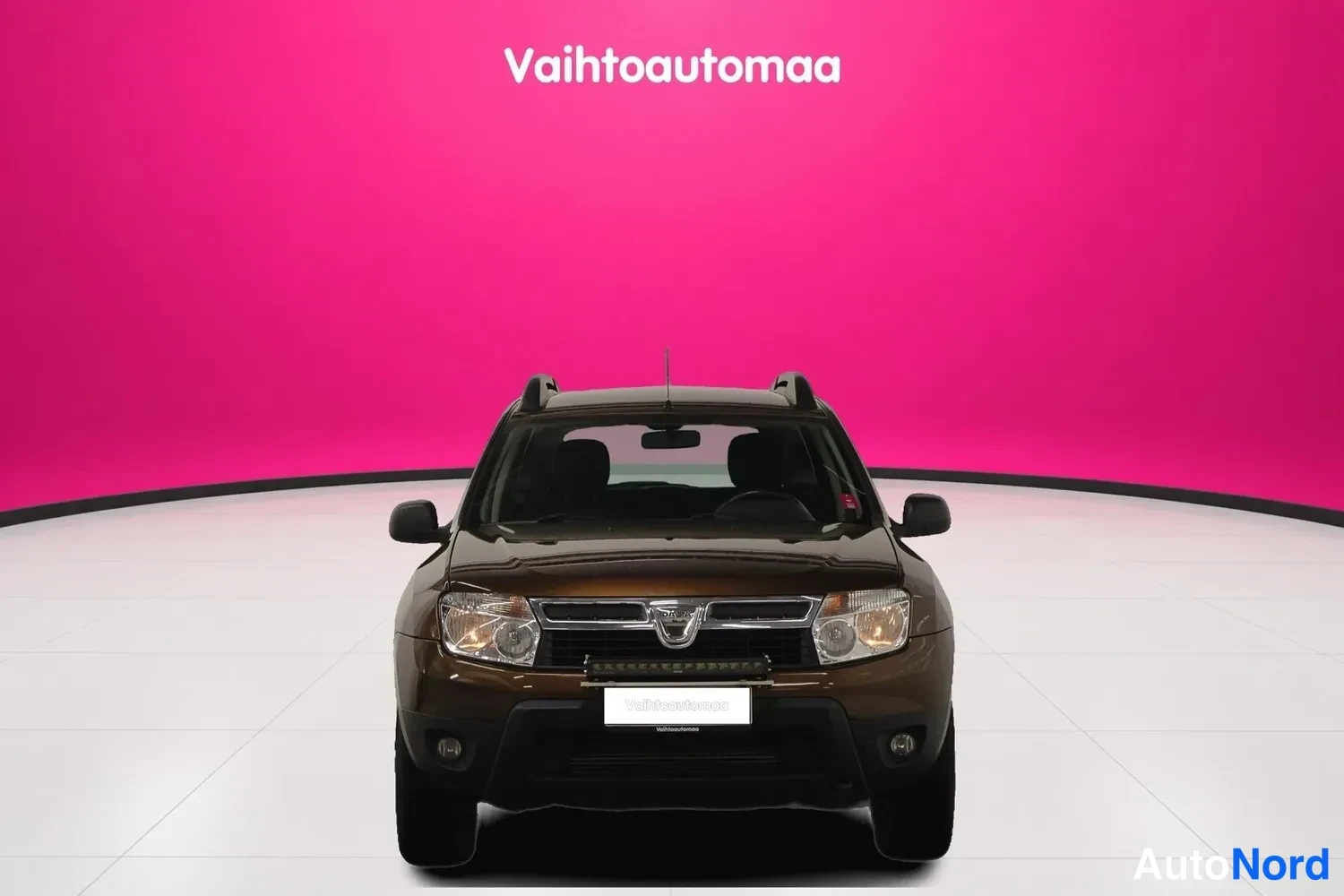 Edestä: Ruskeanmatleanruskea henkilöauto suoraan edestä, etusäleikkö, ajovalot ja etupuskuri näkyvissä valkoisella studiossa pinkin taustan edessä