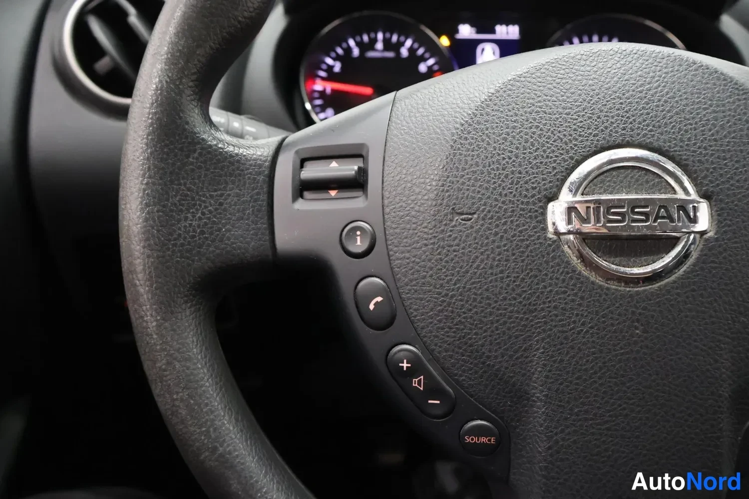 Yksityiskohta: Ohjauspyörän keskusta NISSAN-merkillä ja ohjainpainikkeet, taustalla osa mittaristosta.