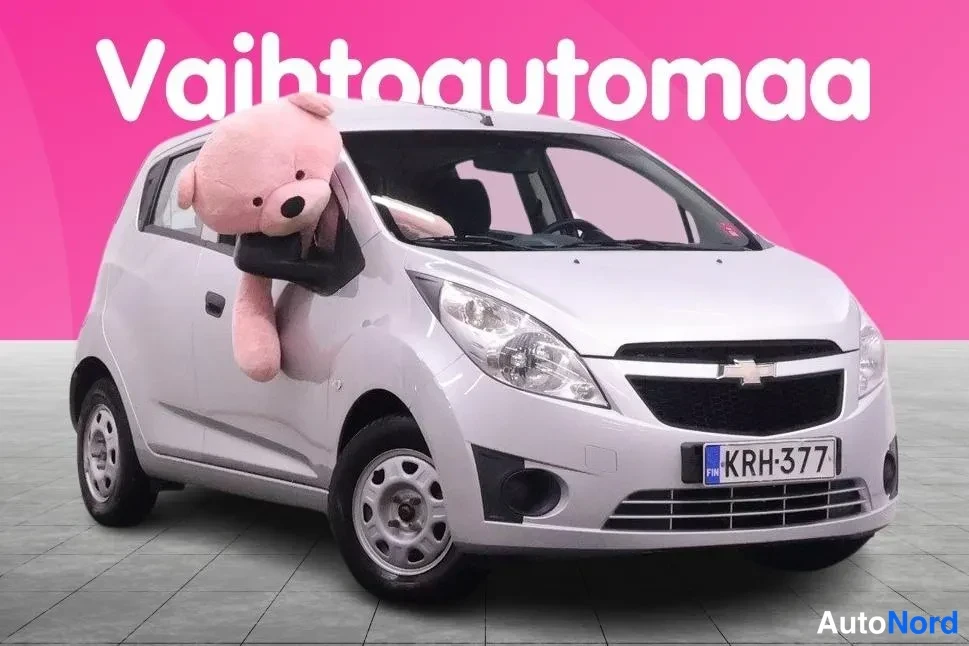 Etuviisto, nokka oikealle: Hopeanvärinen pieni automerkki Chevrolet Spark etuviistöstä oikealta, peilistä roikkuu pehmolelu.