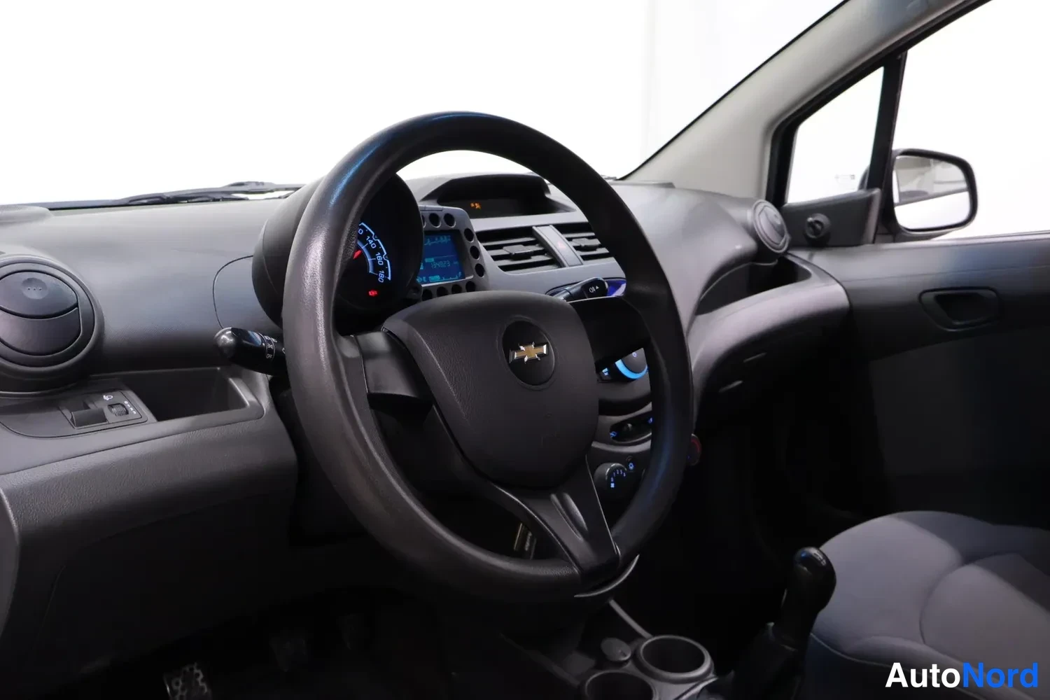 Muu: Kuljettajan näkökulma kojelautaan: Chevrolet-merkitty ohjauspyörä, analoginen nopeusmittari ja manuaalivaihtohippa.