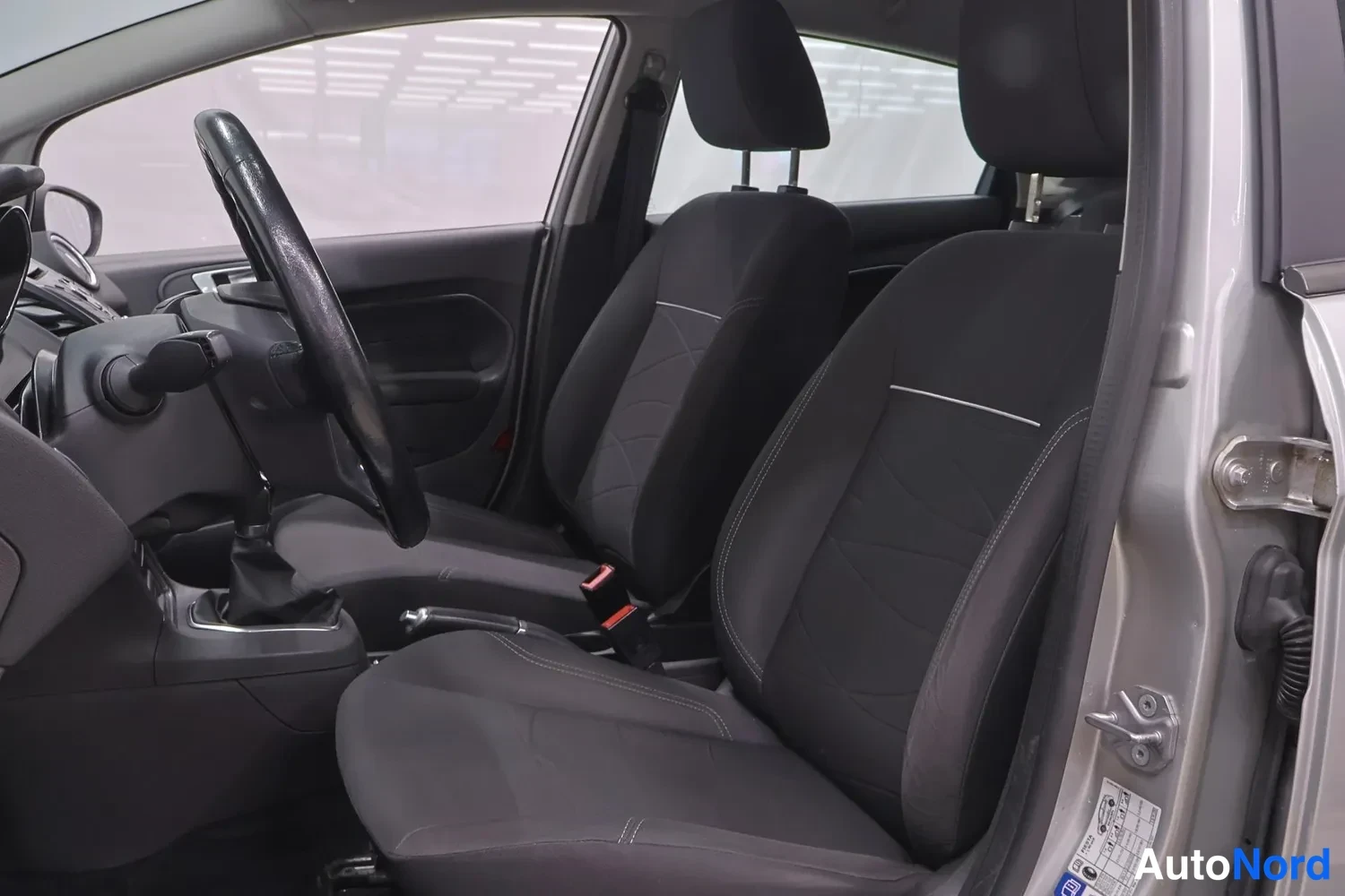 Muu: Ford Fiestan etumatkustamo, kaksi tummaa kangasistuinta, manuaalivaihteen keksi näkyvissä.