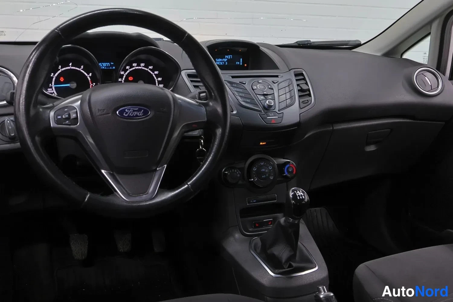 Muu: Kuljettajan puoleinen näkymä: Ford-ohjauspyörä, mittaristo ja manuaalivaihteen keppi, musta kojelauta.