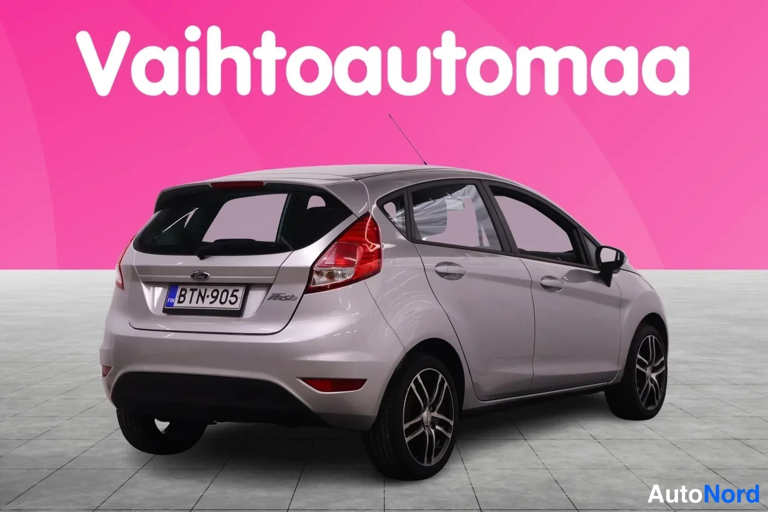 Takaviisto: Hopeanharmaa Ford Fiesta takaa vinosti kuvattuna, rekisterikilpi, vaaleanpunainen studio-tausta.