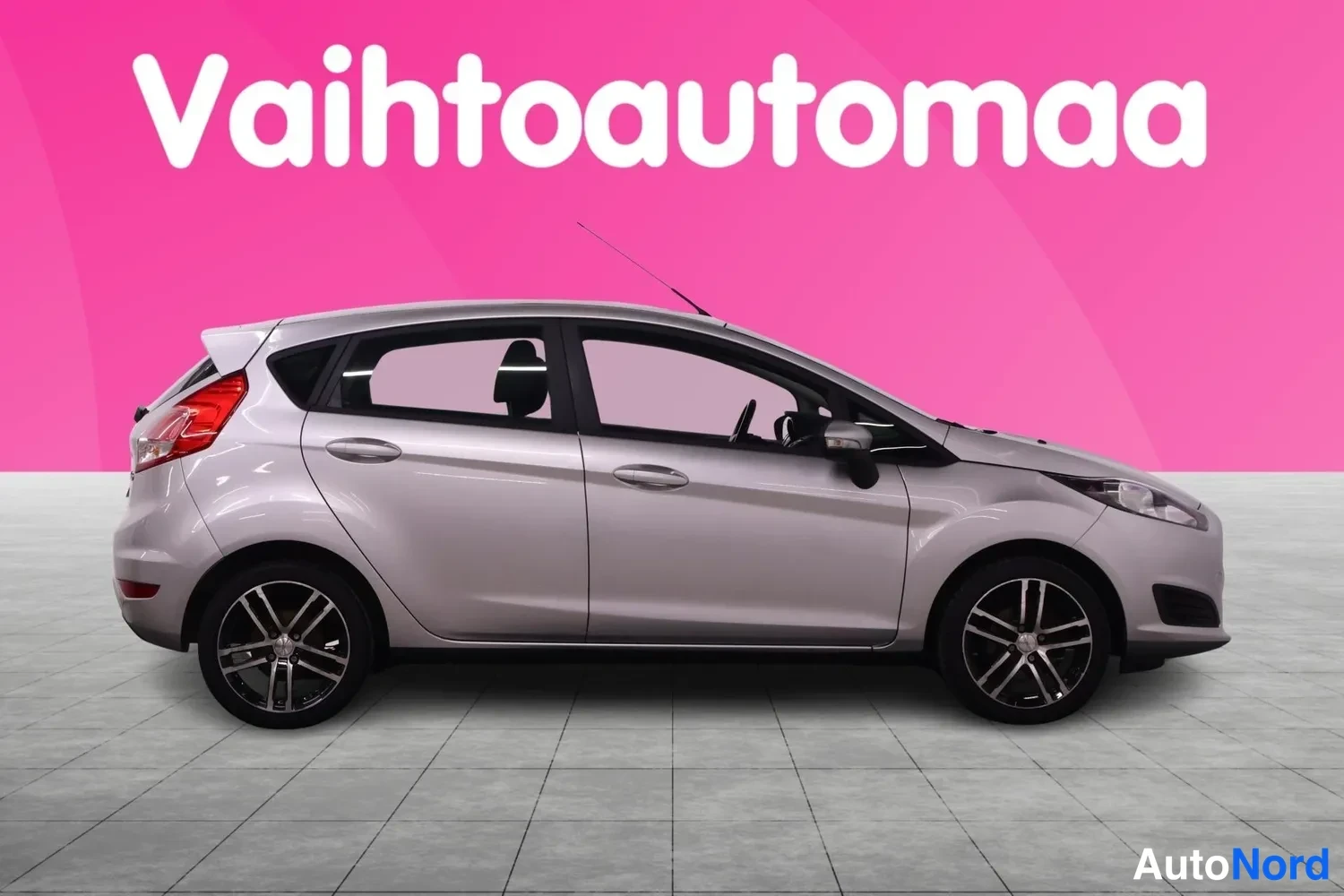 Etuviisto, nokka oikealle: Hopeanharmaa Ford Fiesta sivulta vinosti etukeula oikealla, viisiovinen malli, vaaleanpunainen studio-tausta.