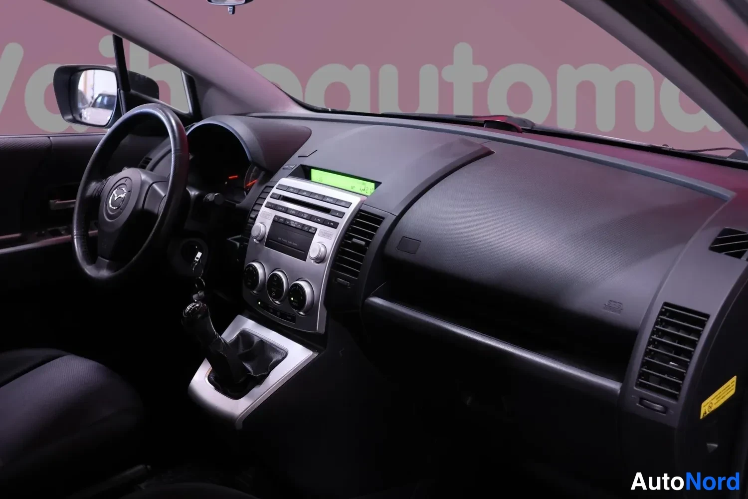 Muu: Kuljettajan näkökulma Mazda 5:n ohjaamoon: tumma kojelauta, radio- ja ilmastointilaitteet sekä manuaalivaihteen vaihdekeppi.