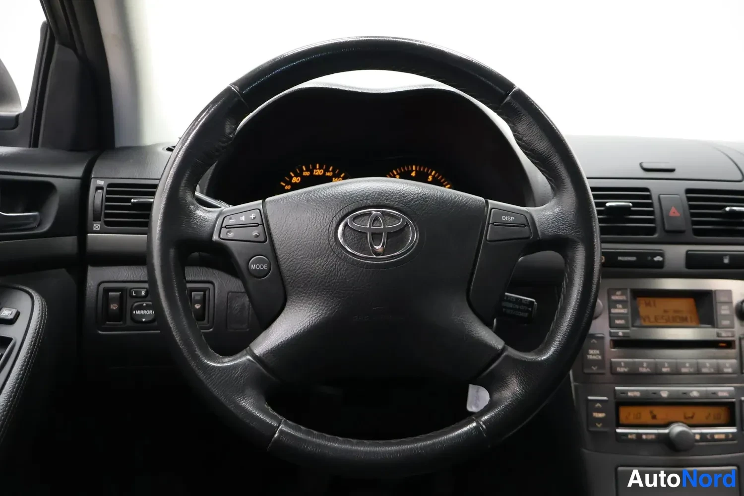 Muu: Ohjauspyörä suoraan edestä: musta ohjauspyörä Toyotan merkillä, oranssit mittarit taustalla ja keskikonsolissa radio sekä lisätasoite-/ajotietonäyttö.