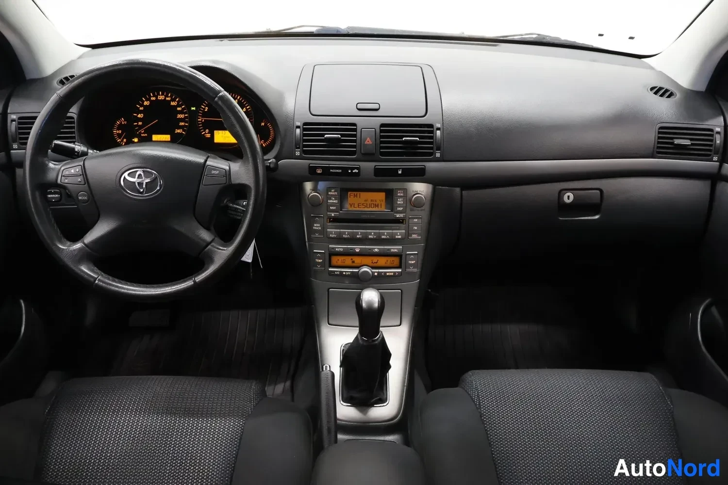 Muu: Kuljettajan näkökulma kojelautaan: Toyota-ohjauspyörä, oranssit mittarit, keskikonsolin radio- jailmastointiyksikkö, manuaalivaihtopyörä, parkkiharausjarru ja e