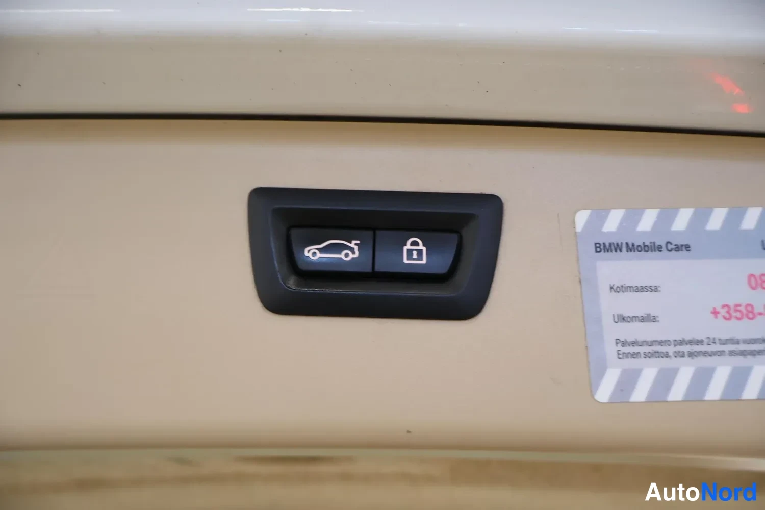 Yksityiskohta: Yksityiskuva bensiinin täyttöaukon luukusta: pieni tumma paneeli, jossa kaksi nappia — avaus ja lukko. Oikealla tarra BMW Mobile Care.