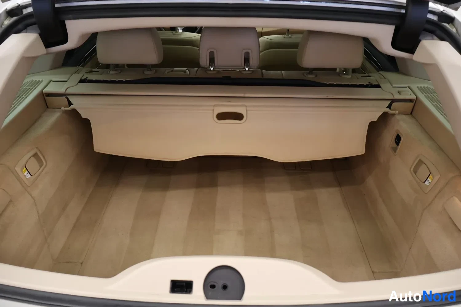 Tavaratila: View of the trunk with a beige retractable cargo cover in place, showing three rear headrests behind it and carpeted floor.