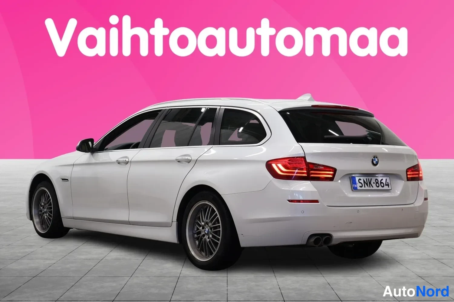 Takaviisto: BMW 5-sarjan farmariautu takaa sivulta, valkoinen, rekisteritunnus, kaksoispakoputki ja kiinteät kattokaiteet näkyvät.