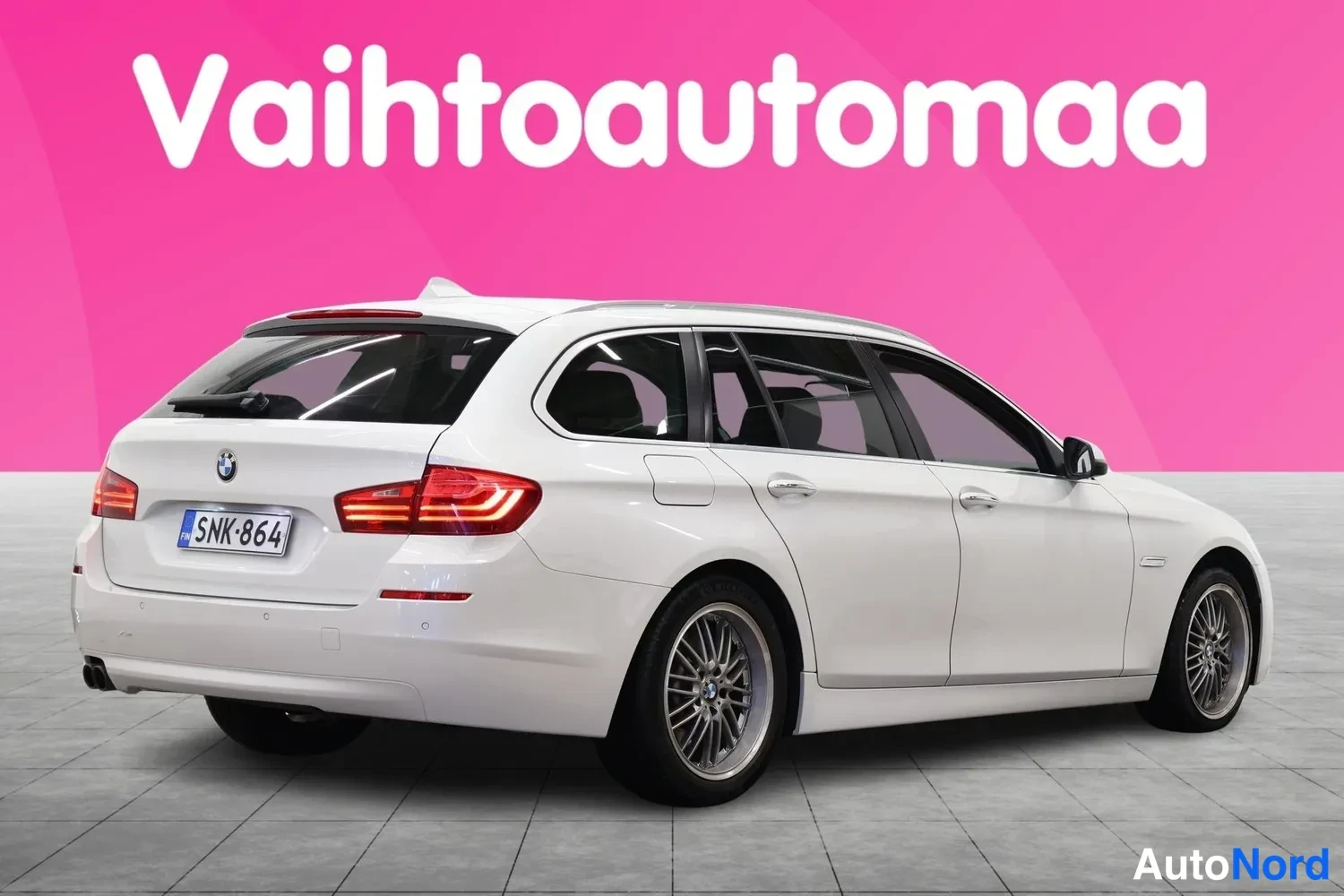 Takaviisto: BMW 5-sarjan farmariautu takaa sivulta, valkoinen, rekisteritunnus, kiinteät kattokaiteet ja kaksoispakoputki näkyvät.