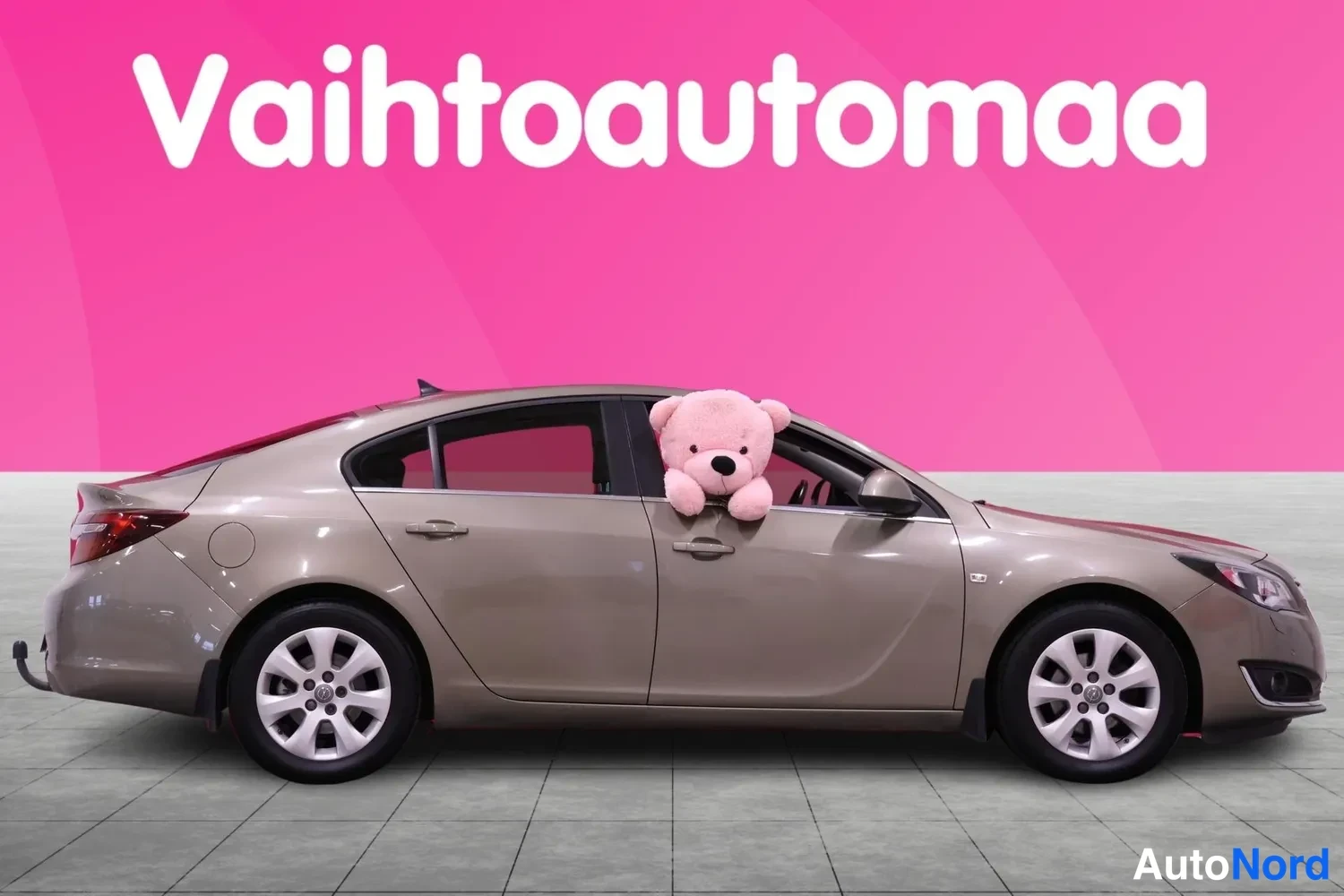 Sivulta: Ruskeanharmaan sedan-auton sivuprofiili vasemmalta, auton sisällä vaaleanpunainen pehmolelu.