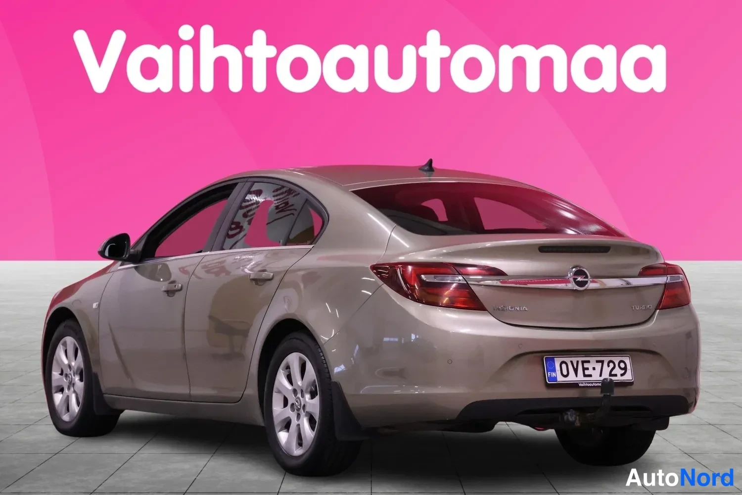 Takaviisto: Used car photographed from the rear three-quarter angle, rear face and left flank visible, beige paint, showroom background.