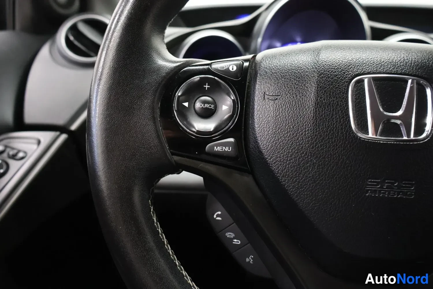 Yksityiskohta: Musta Honda-ohjauspyörä, jossa SRS AIRBAG -merkintä, SOURCE- ja MENU-painikkeet sekä puhelin- ja ääniohjauspainikkeet.