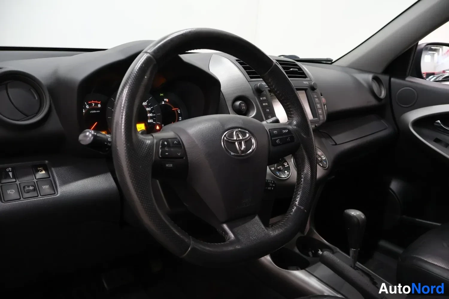 Muu: Kuljettajan näkymä Toyota-merkillä varustettuun ohjauspyörään ja kojelautaan, mittaristo ja äänentoisto- ja navigointiyksikkö näkyvissä.