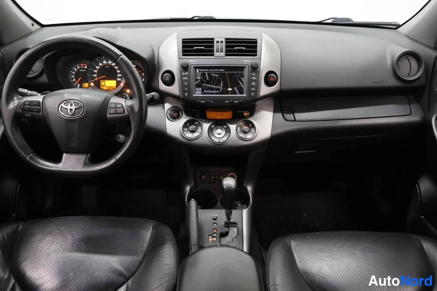 Muu: Ohjaamo kuvattuna kojelautaan päin: Toyota-ohjauspyörä, mittaristo ja keskikonsoli, jossa kosketusnäyttö näyttää karttaa ja teksti 'Bluetooth connection failed'