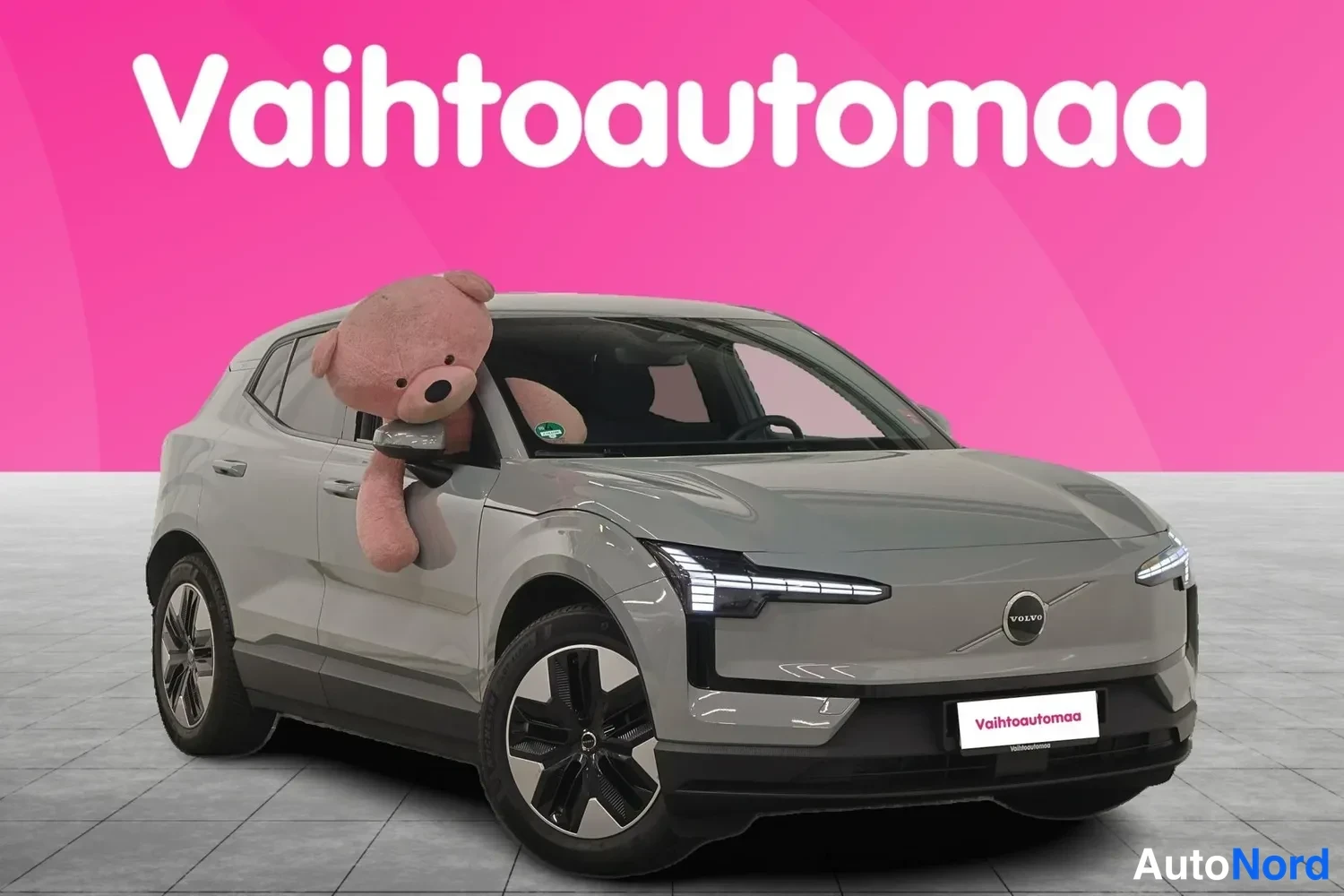 Etuviisto, nokka oikealle: Auton etukulmakuva vasemmalta: harmaanharmaa SUV-sta kappaletta edessä ja kyljessä, A-pilarissa suuri ruskeanpunainen pehmoeläin, tausta pinkki.
