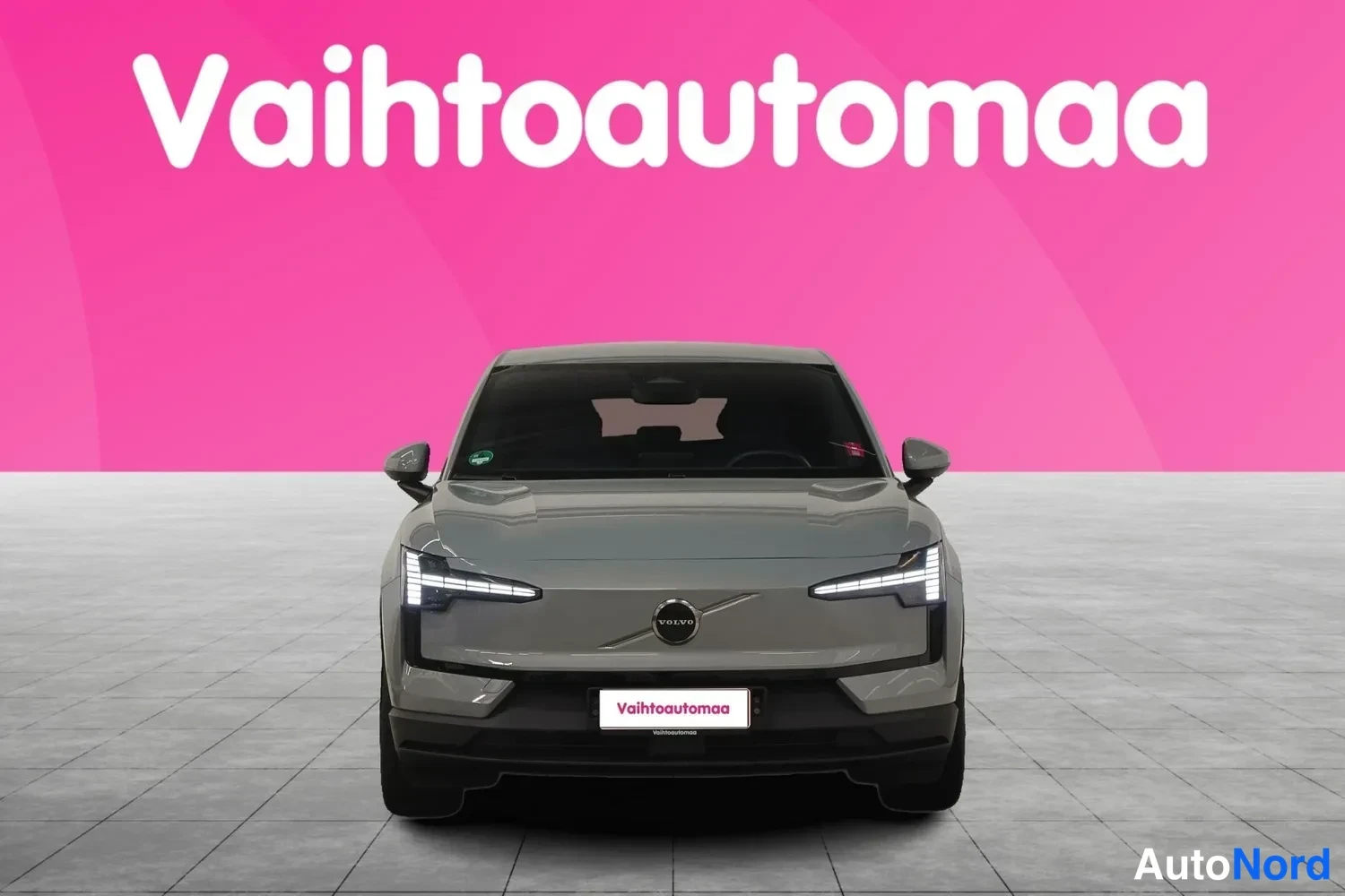 Edestä: Auton suora etunäkymä: harmaanharmaa kori,Volvo-merkki konepellillä ja neonedut valot, tausta pinkki.