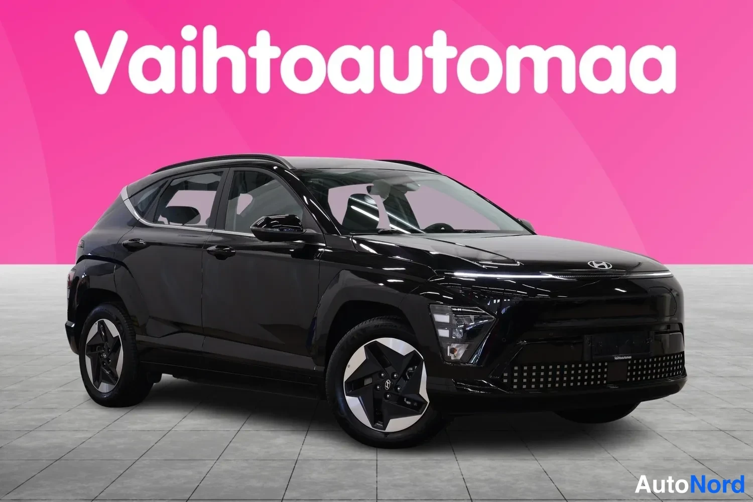 Etuviisto, nokka oikealle: Mustan Hyundai Kona -sähköauton etukulmakuva, keula ja vasen sivu näkyvissä pinkkiä taustaa vasten.