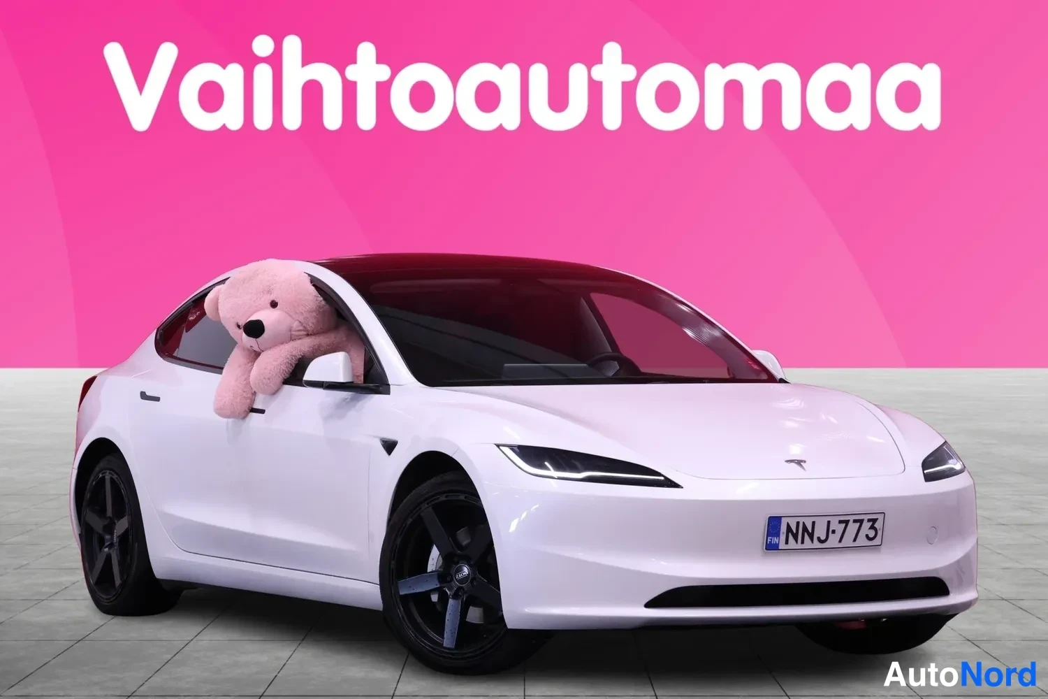 Etuviisto, nokka oikealle: Valkoinen Tesla Model 3 -henkilöauto vinosti edestä oikealta, rekisterilla, kuljettajan ikkunassa pehmolelu.