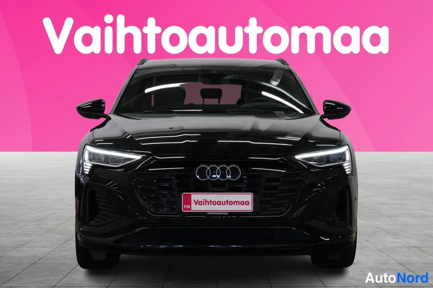 Edestä: Mustan Audin suora etunäkymä, nelirenkaat-maski ja Vaihtoautomaan rekisterikilpitunniste, vaalea hallilattia ja pinkki tausta.