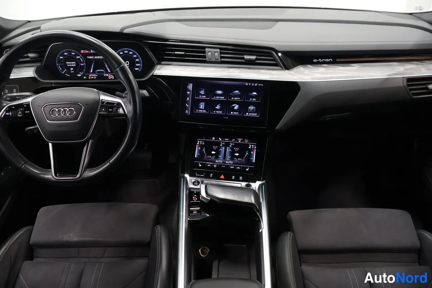 Sivulta: Auton ohjaamo etukädestä: musta sisusta, Audi-merkkinen ohjauspyörä, digitaalinen mittaristo, vaakasuora keskinäyttö ja ilmastointipaneeli (21 °C, AUTO); keskik