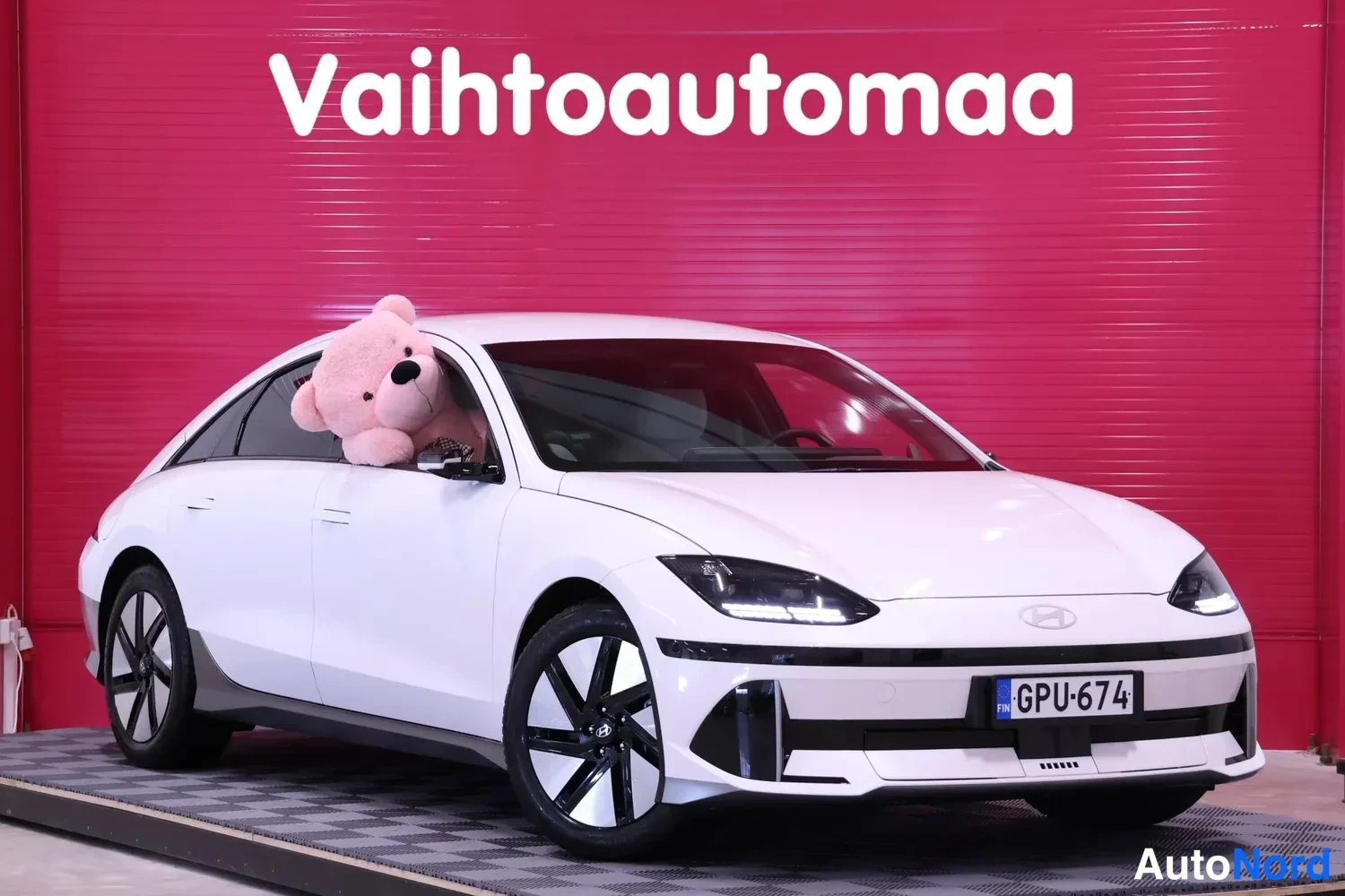 Etuviisto, nokka oikealle: Valkoinen Hyundai Ioniq 6 etu kolmannes oikealta, suuri vaaleanpunainen pehmolelu takapenkillä ikkunan takana, rekisteri, punainen tausta.