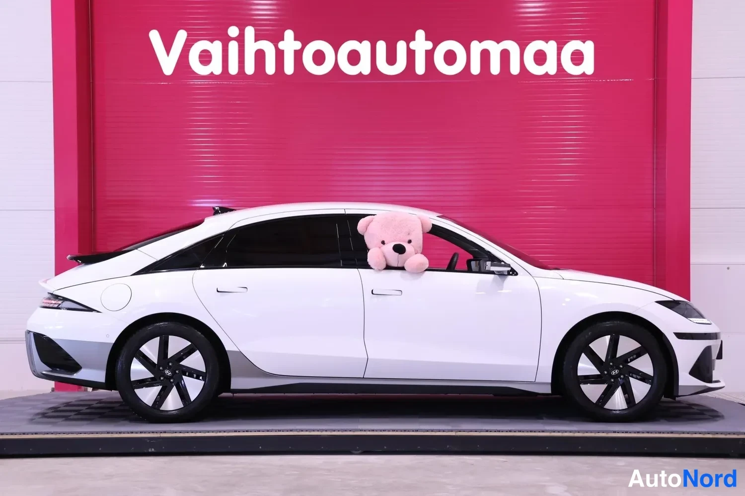 Sivulta: Valkoinen Hyundai Ioniq 6 sivulta oikea kylki näkyvissä, pehmolelu etuikkunan takana, punainen tausta.