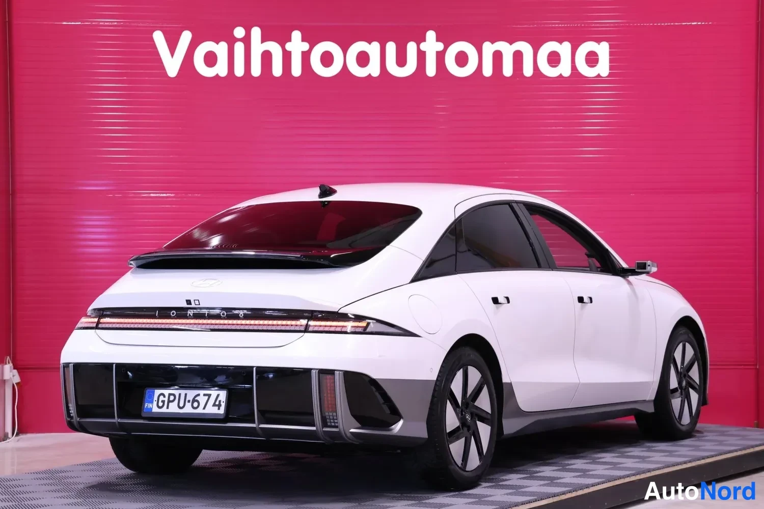 Takaviisto: Valkoinen Hyundai Ioniq 6 takakolmannes oikealta, vaaka-takavalot, rekisteri, punainen tausta.