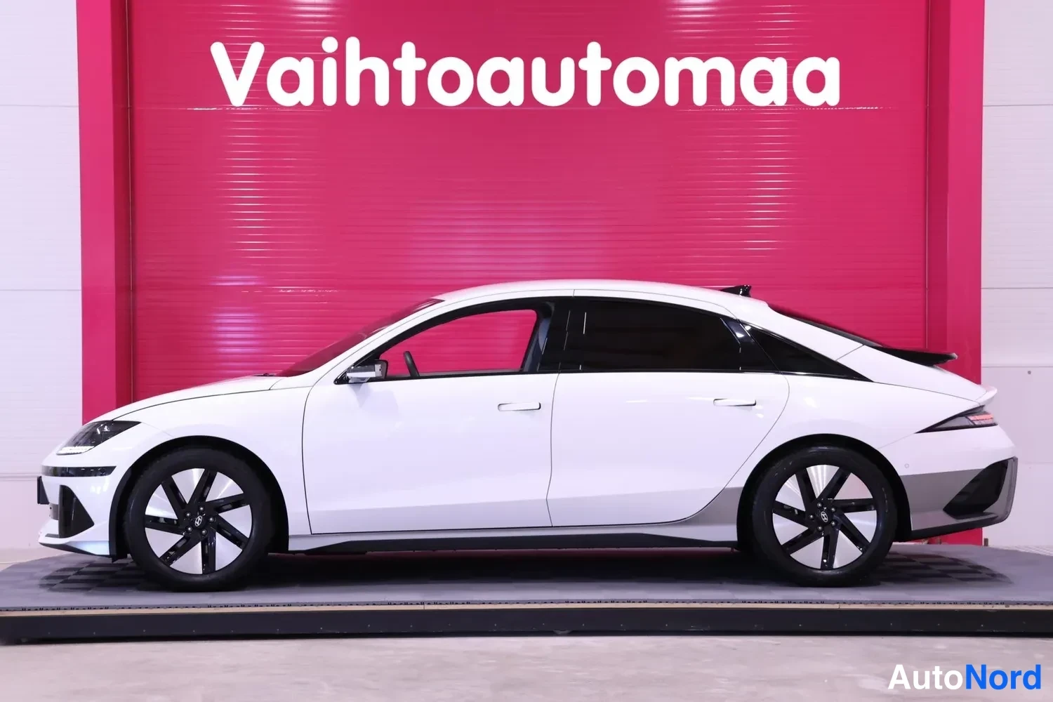 Etuviisto, nokka vasemmalle: Auton kylki ja keulan kärki kuvan vasemmassa reunassa, valkoinen korinpinta, tummat ikkunat.