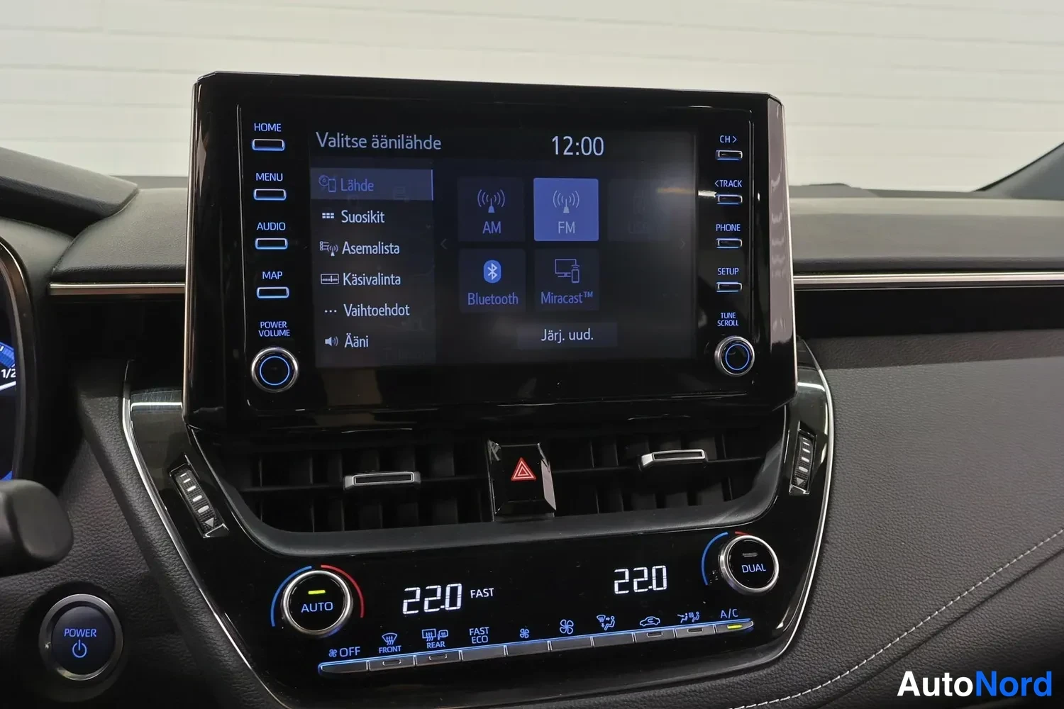 Muu: Keskikonsolin infotainment-näyttö valikossa 'Valitse äänilähde', jossa vaihtoehdot AM, FM, Bluetooth ja Miracast; kellonaika 12:00; alla ilmastointipaneeli 22.0