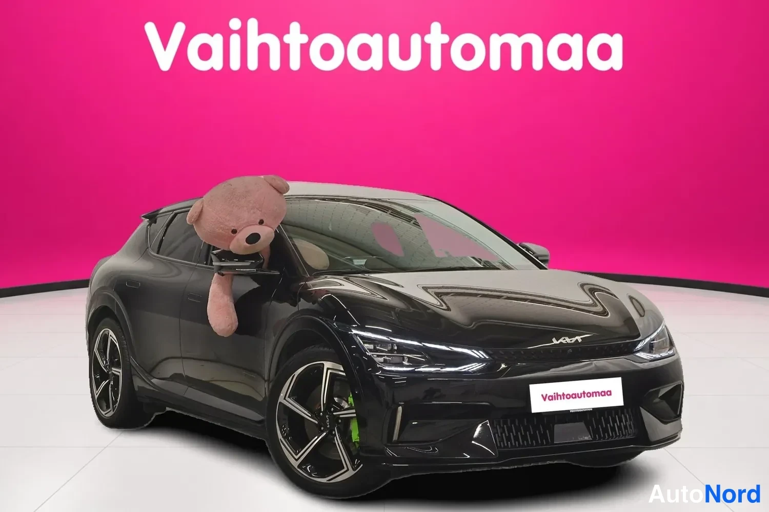Etuviisto, nokka oikealle: Musta Kia EV6 etukulmasta oikealta, pehmoauto ikkunassa, Vahtoautomaan taustalla.