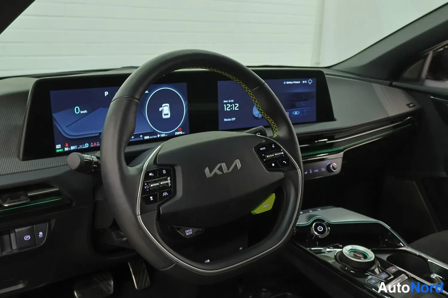Muu: Ohjauspyörä, jossa on Kia-logo, ja vihreä tikitys yläreunassa. Digitaalinen mittaristo näyttää 0 km/t ja P-vaihteen, keskustietonäyttö näyttää kellon 12:12. Kon