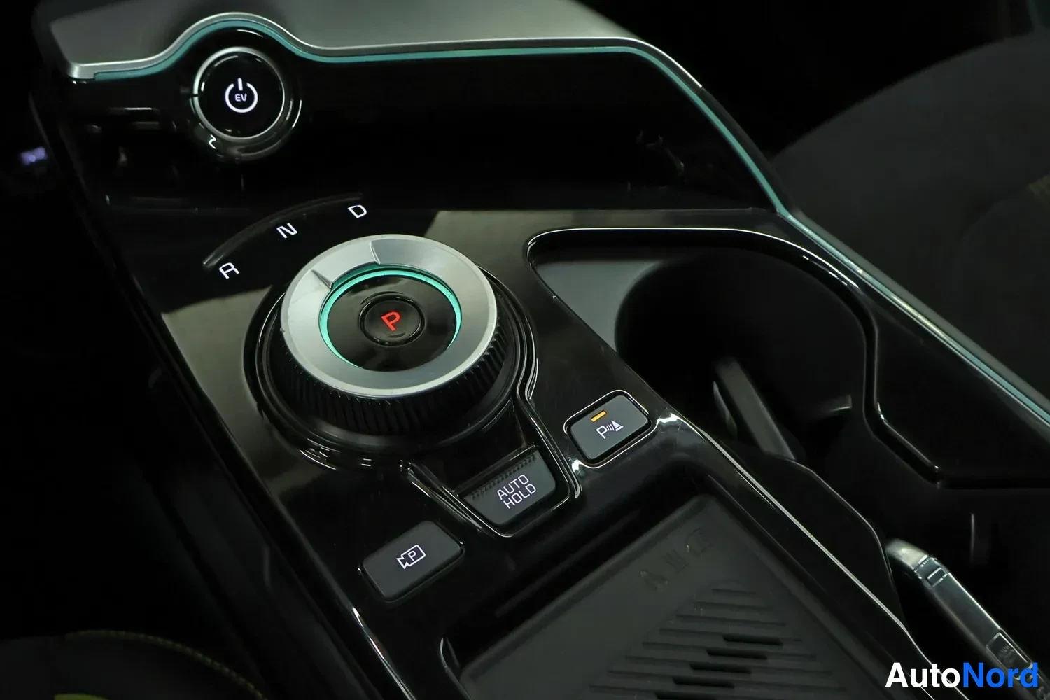 Yksityiskohta: Close-up view of the car's center console gear selector dial with 'P R N D' letters, electronic parking brake button with 'P' and parking sensor symbol, 'AUTO H
