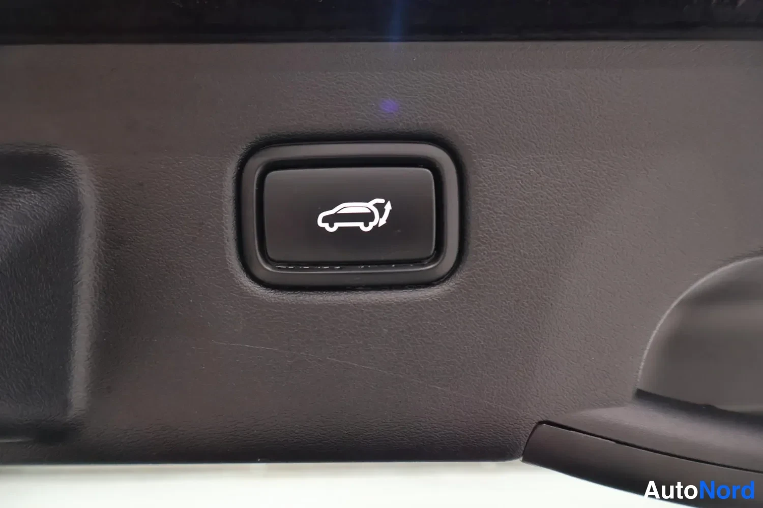 Yksityiskohta: Interior switch panel detail showing a raised rectangular button with a white icon depicting a hatchback with its rear door open