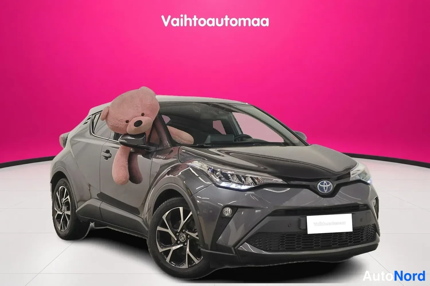 Etuviisto, nokka oikealle: Tummanharmaa Toyota C-HR esitettynä etuosa vasemmasta kulmasta, suuri pehmolelu takana.