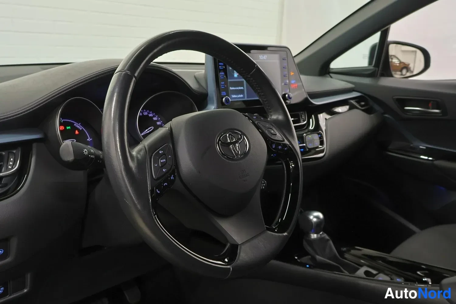 Muu: Ohjaamo etupuolelta: Toyota-merkitty ohjauspyörä, digitaalinen mittaristo ja keskinäyttö, vaihteenvalitsin näkyvissä.