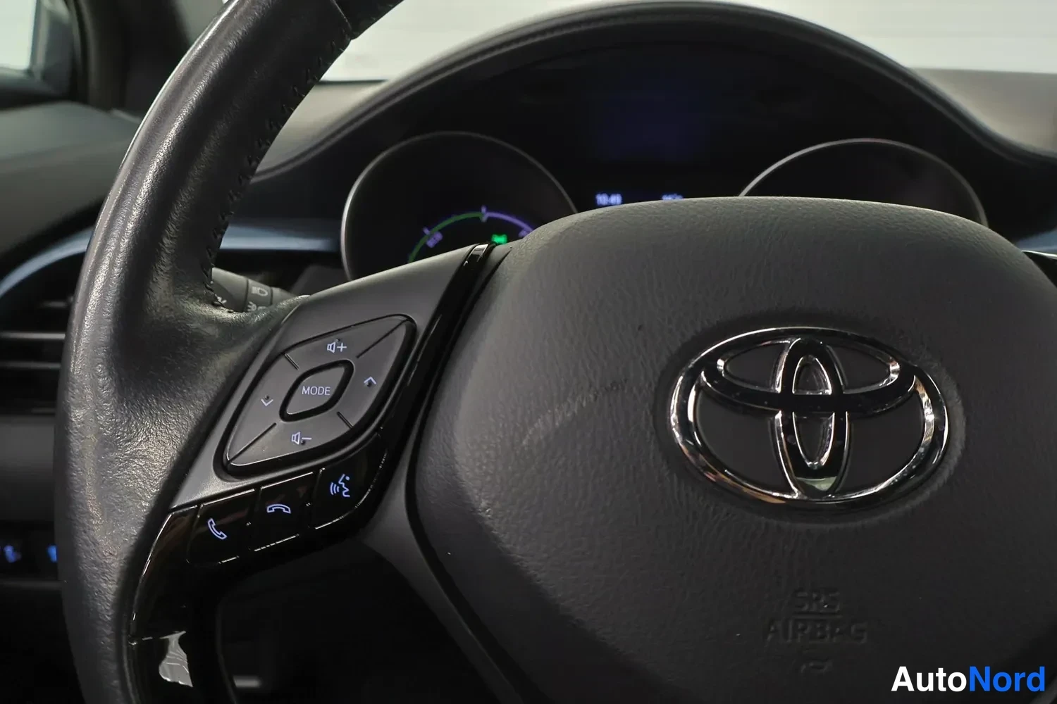 Yksityiskohta: Rattilähikuva: Toyota-merkki keskellä, vasemmassa puhelin- ja audio-ohjaimissa MODE- ja puhelinnäppäimet, taustalla mittaristo.