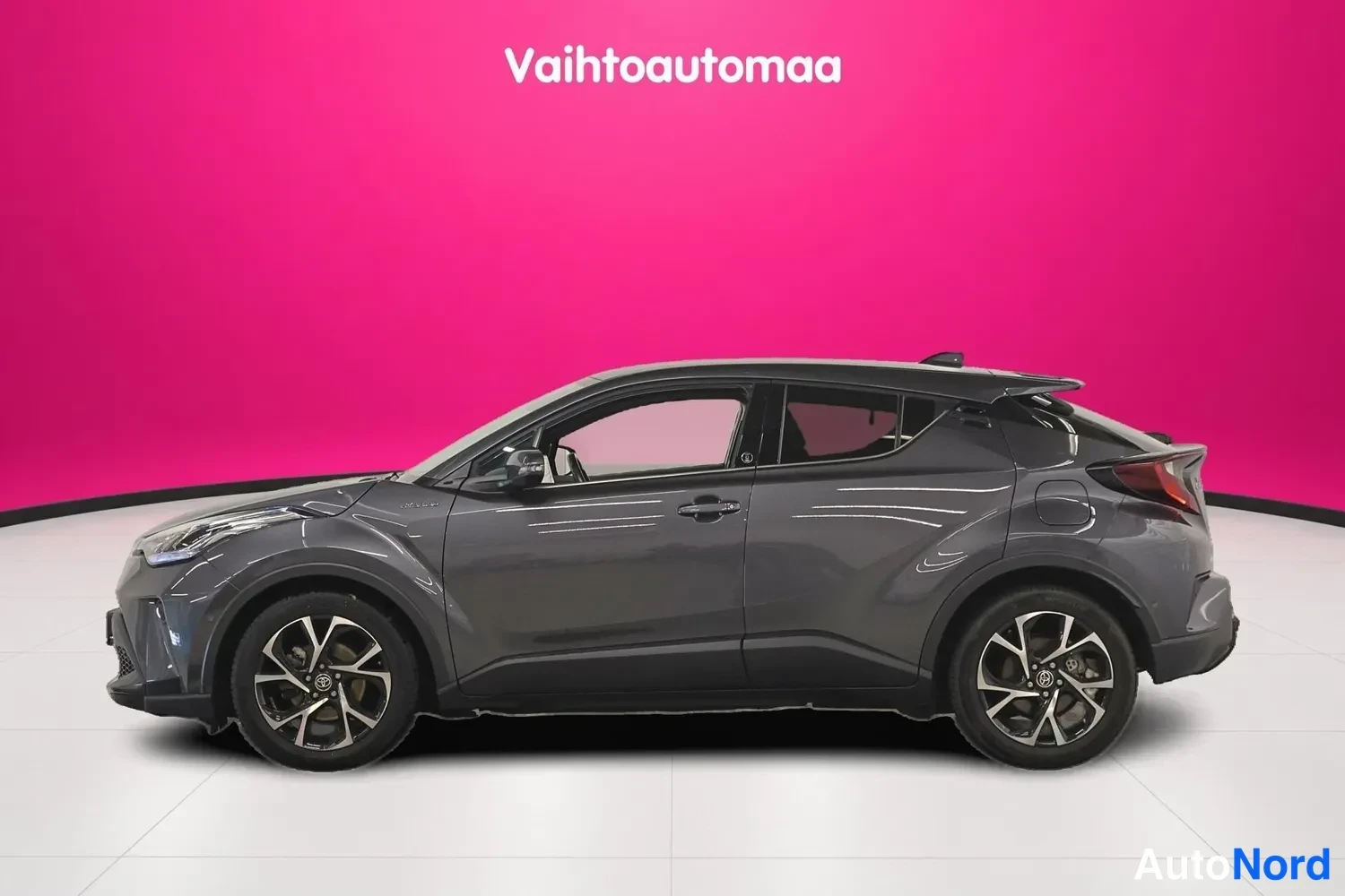 Etuviisto, nokka vasemmalle: Tummanharmaa Toyota C-HR vasemmalta sivulta etukulmasta, keula vasemmalla, tummanpunainen tausta.