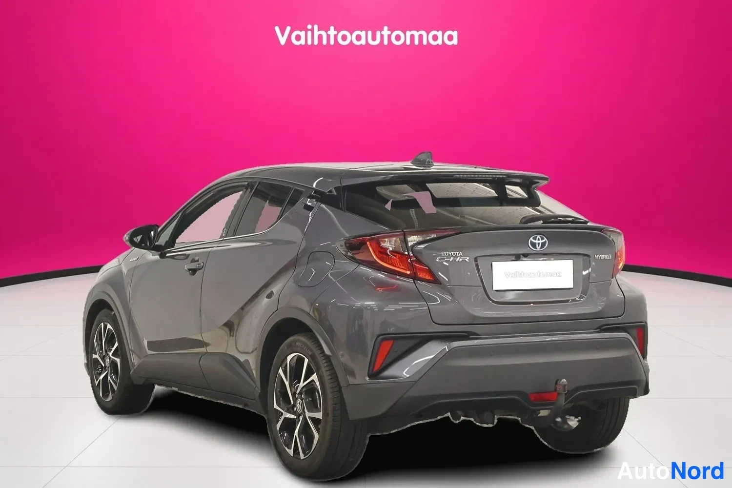 Takaviisto: Tummanharmaa Toyota C-HR takakulmasta, perä ja vasen sivu näkyvissä, tummanpunainen tausta.