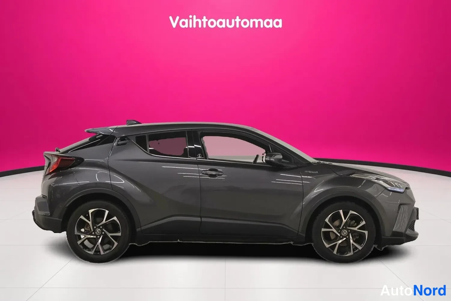 Etuviisto, nokka oikealle: Auton sivuprofiili ja keula vinosta etunurkkakulmasta oikealta: tummanharmaa Toyota C-HR, hybrid-merkintä etulokasuojassa.