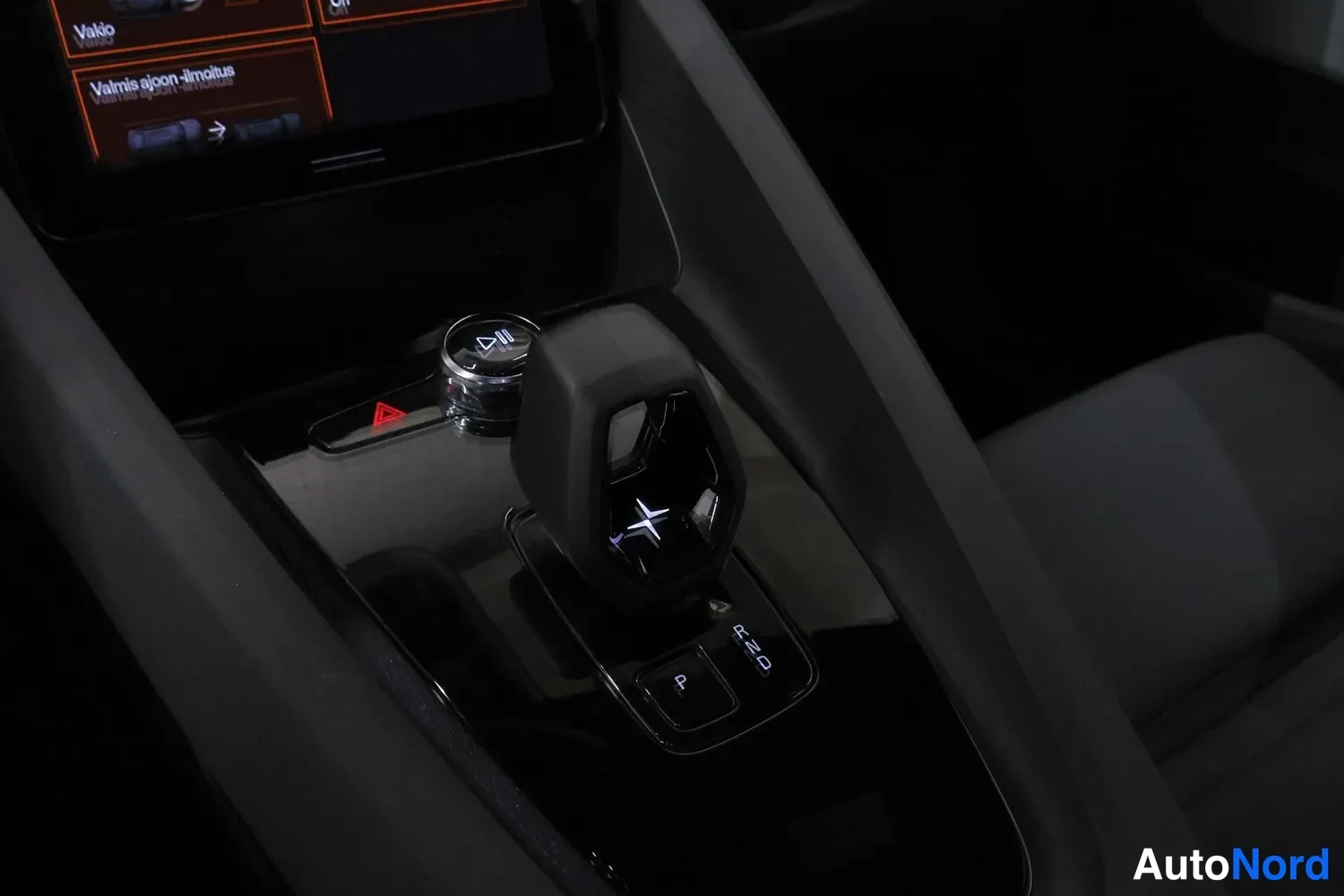 Yksityiskohta: Close-up of the center console with a short gear selector featuring the Polestar logo. An illuminated start/stop button and control buttons are visible on the g