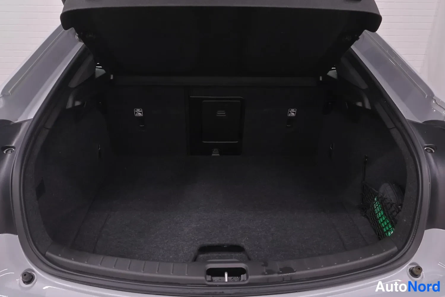 Tavaratila: Rear cargo area of an electric vehicle with the hatch fully open. The trunk is lined in dark grey carpeting and features a storage net on the right interior sid