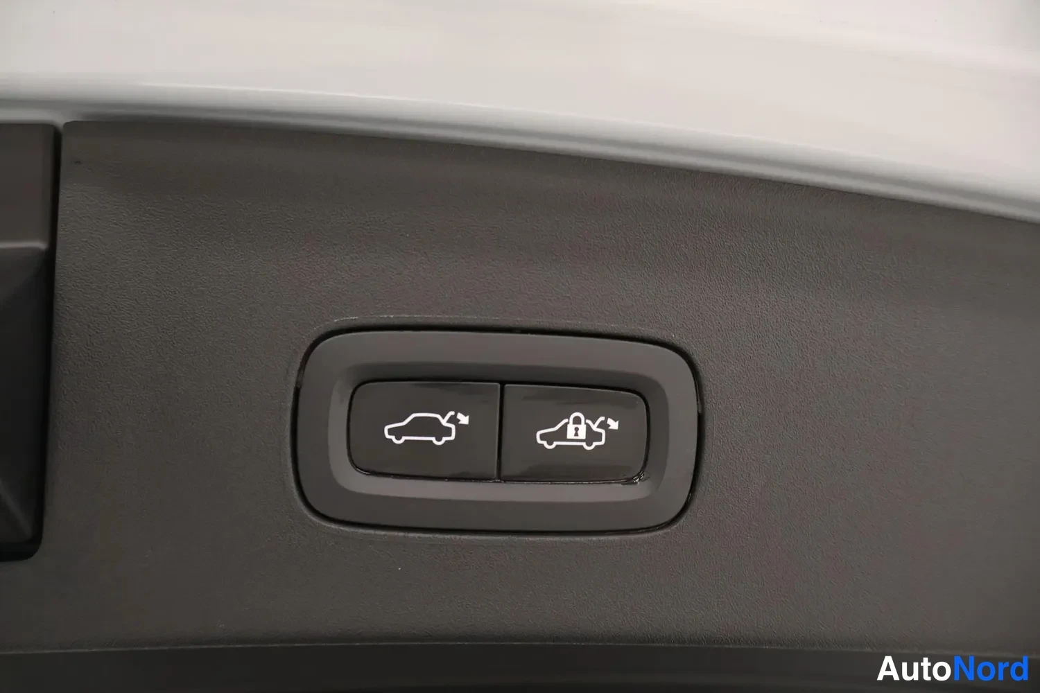 Yksityiskohta: Detail view of two buttons mounted on a black plastic trim piece inside the boot. The left button displays a car icon with an open trunk (tailgate release), and