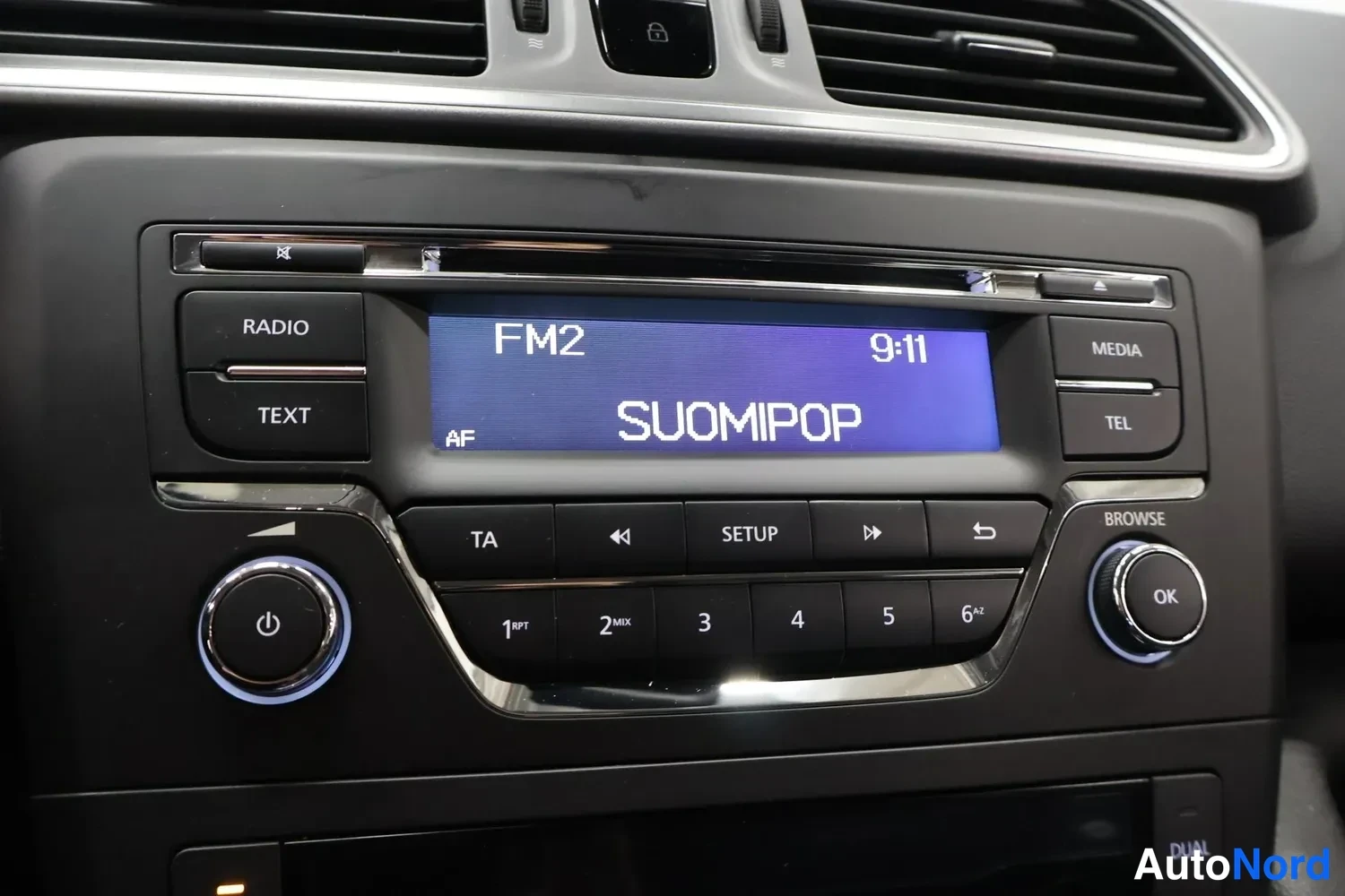 Yksityiskohta: Autoradion paneeli: näytöllä FM2, kello 9:11 ja teksti SUOMIPOP; painikkeet RADIO, TEXT, MEDIA, TEL, TA, SETUP ja numeronäppäimet.