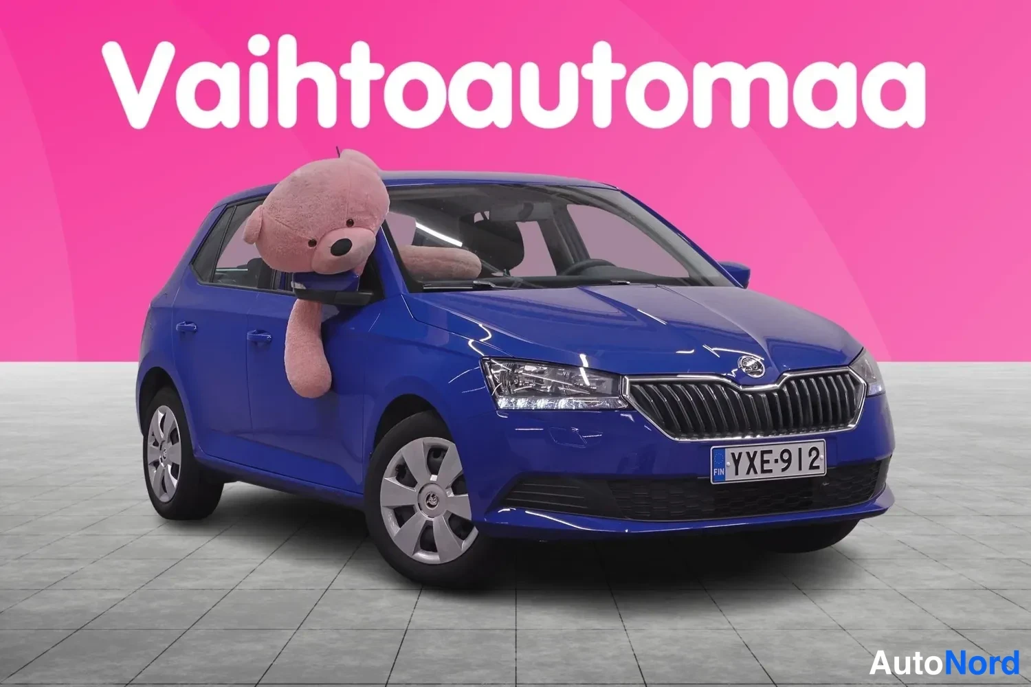 Etuviisto, nokka oikealle: Sininen Skoda Fabia etukulmasta oikealta, rekisteritunnus, vaaleansininen nallekeinu ikkunassa Vahtoautomaan markkinointitaustaa vasten.