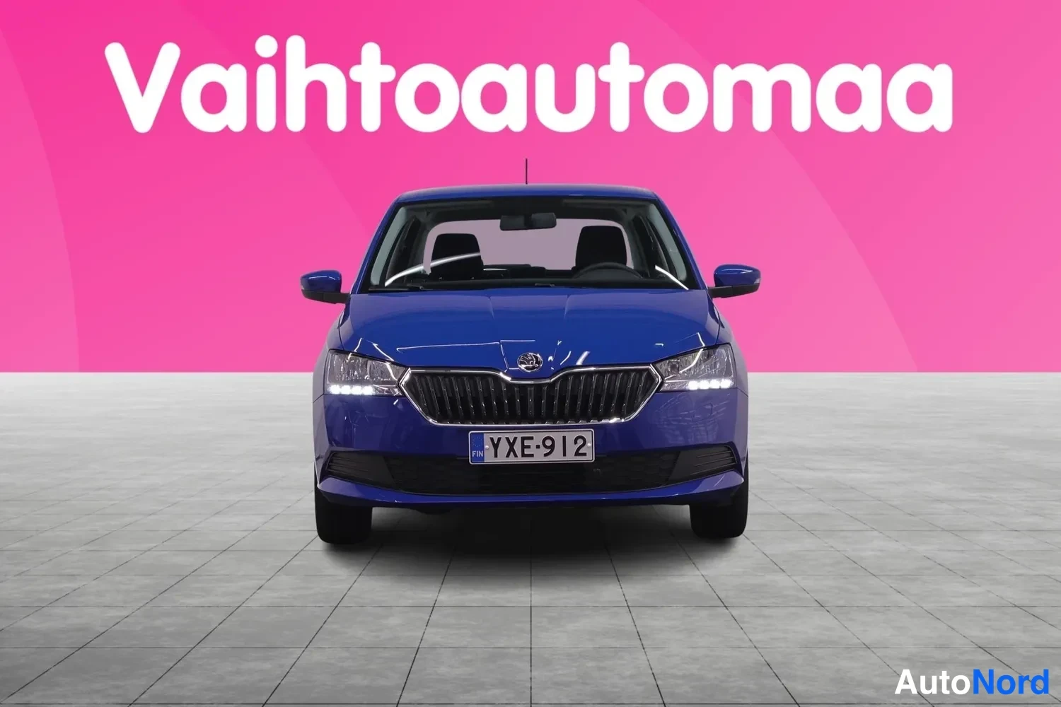 Edestä: Sininen Skoda Fabia suoraan edestäpäin, rekisteritunnus, Vahtoautomaan markkinointitaustaa vasten.